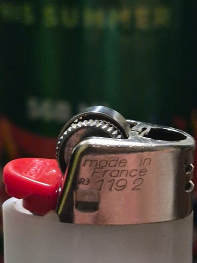 The image shows a close-up of a BIC lighter. The lighter is metallic with a red plastic cap. Text on the lighter reads "made in France" and "119 2". The background is out of focus and appears to be dark green with some blurry yellow shapes, possibly text. There is no indication of a specific location in Sooblase, Estonia, in this image.