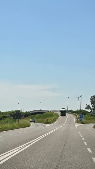 The image shows a road in San Michele al Tagliamento, Italy. The road is paved and has white lines indicating lanes. There is a green truck driving up a hill on the right side of the road. A car is driving on the left side of the road. There is a bridge in the background and trees and grass on the sides of the road. The sky is blue with a few clouds. There are several street lights visible. There are also road signs with directional information and symbols. The overall scene suggests a highway or a major road in a rural or suburban area.