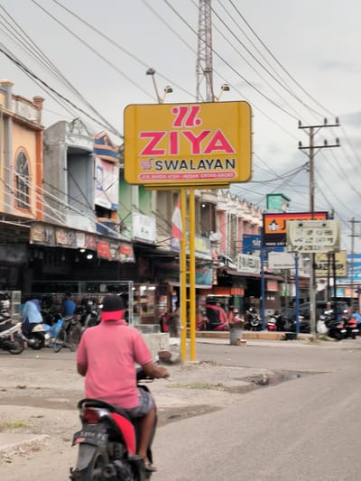 An outdoor, eye-level shot captures a bustling commercial street scene in Grong-Grong, Indonesia, under an overcast sky. The image is dominated by a large, bright yellow sign for "ZIYA SWALAYAN" in the mid-ground, featuring red lettering and a shopping cart icon. Below the main name, smaller text reads "JL. BANDA ACEH - MEDAN, GRONG-GRONG," providing a clear location cue.

In the foreground, a person wearing a pink shirt and a red cap rides a red and black scooter away from the viewer, its license plate "5819 PE" partially visible. The street surface is asphalt, with some dirt and gravel on the shoulders. Numerous other motorcycles and scooters are visible, parked or in motion, indicating a common mode of transport in the area.

The background features a row of multi-story buildings, housing various shops or businesses, with diverse architectural styles and colors, including orange, grey, and green facades. Overhanging awnings or display racks are visible outside some establishments. A dense network of power lines and communication cables crisscrosses the sky above the street, connected to wooden power poles on the right and a tall metal tower on the left. Other, smaller commercial signs are scattered along the street, though mostly blurred or illegible, with some partial text discernable as "sir," "TOED CAT," and "MUUND SHIMAT." The overall scene depicts a typical busy daytime street in an Indonesian town.