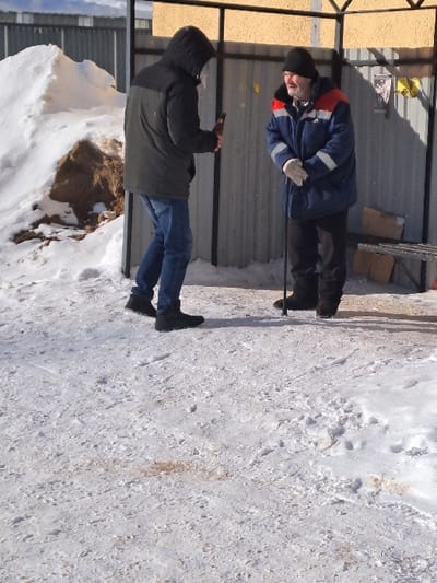 The image captures an outdoor scene in Bolshaya Elhovka, Russia, on a sunlit winter day. The ground is covered with a thin, irregular layer of snow and ice, revealing patches of brown dirt.
Two adult males are present. The individual on the left, positioned with his back partially towards the viewer, wears a dark, two-tone hooded winter jacket, blue jeans, and dark boots. He holds a dark glass bottle in his right hand at waist height.
The individual on the right, an older male with a beard, faces the first man. He wears a dark blue winter jacket with red and white reflective stripes on the chest and sleeves, a black knit cap, black trousers, and light-colored gloves. He holds a dark walking stick or cane vertically in front of him with both gloved hands. He stands beneath a black metal framework structure, behind which a corrugated metal fence and a yellow textured wall are visible. Cardboard boxes are stacked on a low surface to his right, against the fence.
To the far left of the frame, a large mound of snow with exposed brown soil or sand is visible. Shadows from the men and the metal structure are cast on the snow-covered ground.