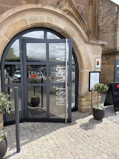 The image shows the entrance to a restaurant in Bad Sobernheim, Germany. The entrance is characterized by a large, rounded, arched opening made of light-colored stone. Within this archway is a modern black framed glass door, consisting of multiple panes. On the glass panes, in large white letters, is the reversed text "RESTAURANT". A logo for "HELZLES. CESAR MORIAN" is visible through the glass, indicating the restaurant's name.

To the right of the door, a paved walkway with small square cobblestones extends. Adjacent to the entrance is a section of the building's exterior, also made of light-colored stone. There is a menu board mounted on the wall. Near the menu board are two large flower pots containing greenery and white flowers. A metal barrier is on the bottom right. A sign saying Standesamt can be seen on the building along with a sign containing the words "Denk" and "Herze We...".