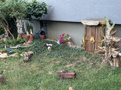 The image depicts a whimsical, outdoor scene that appears to be a decorated yard or garden area. In the upper left, a large tree with lush green foliage overhangs the scene, partially obscuring a light-colored building wall with a horizontal textured pattern. Below the tree, an assortment of garden decorations are visible. These include several carved wooden figures, such as a bear-like creature holding a blue object, a cow with large eyes, and a squirrel. There are also various pots, some containing plants, and a broken terracotta pot. A blue, possibly fabric, creation with a dark opening is partially visible among the greenery.

To the right of the center, a small, rustic wooden door is set into the building's foundation, resembling a miniature entrance. A wooden sign with carved lettering, which appears to read "NARNIA," is mounted above the door. The door is adorned with ivy and a golden lion's head decoration. Beside the door stands a large, weathered, and intricately shaped tree root or stump, also decorated with what appear to be two blue orbs and some leafy elements.

The ground is covered with dense green ivy and patches of grass. Several more decorative items are scattered across the ground, including small wooden animal figures that resemble a dog and a donkey, each with distinct, somewhat comical, facial features. A solar garden light is also visible in the background. The overall impression is one of creative, quirky garden ornamentation. This scene is located in Galanta, Slovakia.