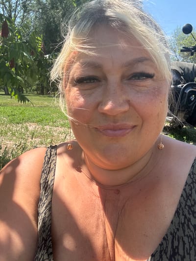 The media file shows a close-up, rotated, selfie-style photo of a person outdoors. The subject has blonde hair and appears to be wearing makeup, including prominent eyelashes. They are looking towards the camera with their eyes open. The background suggests a sunny, outdoor setting with green grass and trees, indicative of daytime. A hint of machinery, possibly a motorcycle or similar vehicle, is visible in the upper left corner. The subject is wearing a patterned top. The scene appears to be set in Zmiiv, Ukraine. The lighting suggests it is a bright, sunny day.