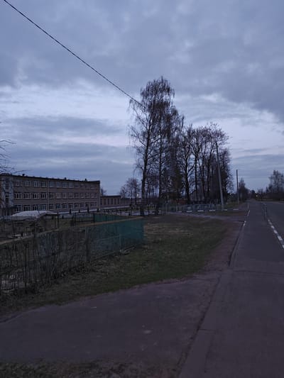 The image depicts an overcast scene in Krychau, Belarus. A multi-story brick building, likely a school or administrative structure, is visible on the left. A green metal fence runs along the foreground. A row of bare trees lines the roadside. A paved road with a painted edge marker extends into the distance. The sky is heavily clouded, suggesting an early morning or late evening.