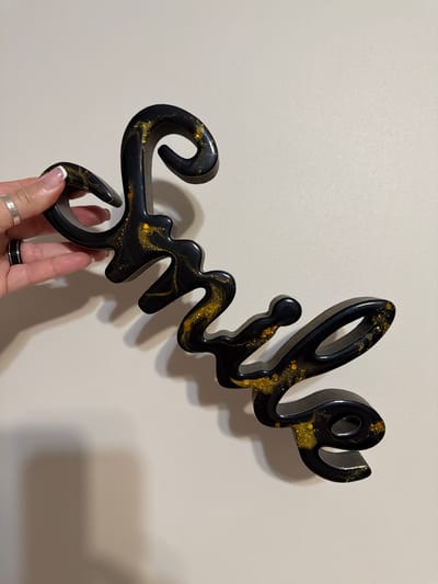 A person's hand, adorned with two rings, is holding a black cursive wall decoration with gold glitter accents. The decoration appears to spell out the word "Smile." The background is a plain, light-colored wall. There are no discernible features or landmarks indicating the specific location within the city of Biržai, Lithuania.