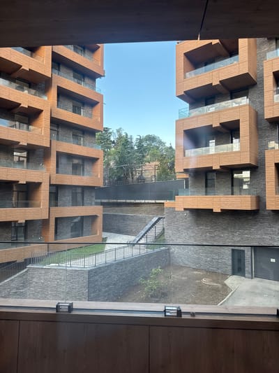 The image shows a view of a modern residential complex, with two buildings visible, one on the left and one on the right. The buildings have a distinctive architectural style, featuring staggered balconies with wood-like cladding and dark stone or brick lower sections. The balconies are equipped with glass railings. The scene appears to be outdoors, with a clear blue sky above. In the middle ground, there are pathways, stairs, and some greenery, suggesting an outdoor courtyard or landscaped area. The foreground is dominated by a wooden interior structure, possibly a balcony or window frame, with a glass barrier. The overall impression is one of contemporary urban architecture. The prompt requested inclusion of the location "Гурзуфский поселковый совет, Ukraine", however, there are no visual elements in the image that confirm this specific location.