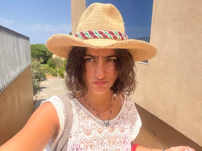 A high-angle, close-up selfie photograph depicts a female individual outdoors in Aljezur, Portugal. The subject, positioned centrally, wears a wide-brimmed straw hat with a red, white, and green band, casting distinctive patterned shadows across her face. Her expression is neutral to slightly somber, with her lips subtly downturned. She has dark, wavy hair and appears to be wearing a white, short-sleeved, open-knit or crocheted top over a pink garment, along with multiple delicate necklaces, including a red-orange beaded strand and a blue beaded strand with a charm. A light grey or tan bag strap is visible over her right shoulder.

The background reveals a narrow outdoor passageway. To the right, a textured, light-colored building wall extends upwards. To the left, a vertical, possibly corrugated or paneled wall is present. A stone-paved path descends into the distance between these structures, flanked by green foliage and mature trees under a clear, bright blue sky. The scene is illuminated by strong sunlight, creating harsh shadows and highlights.