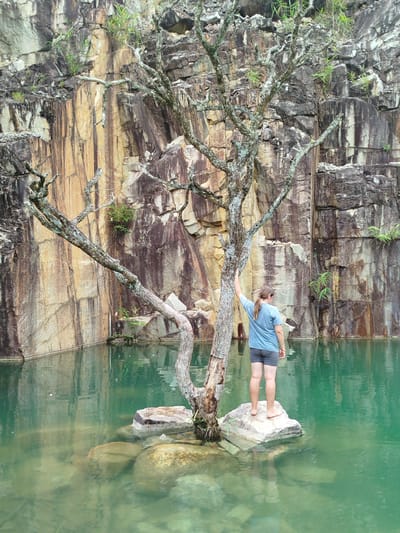 The image depicts a person standing on rocks in the middle of a clear, turquoise body of water. The person is facing away from the camera, wearing a blue t-shirt and grey shorts. They are barefoot and have their left hand resting on a barren tree. The tree's branches are gnarled and bare, with some lichen visible on the bark.
The water is surrounded by steep, sheer rock faces. These rock faces are a mixture of colors, including shades of tan, brown, grey, and white, with visible striations and textures. Patches of green vegetation are growing from crevices in the rock walls. The water reflects the rock faces and the sky, creating a calm and serene atmosphere.
The scene appears to be a quarry or a natural rock formation that has filled with water. The overall impression is one of natural beauty and a sense of isolation. The location is identified as Lán Tranh, Vietnam, suggesting a natural or semi-natural site within the city or its surrounding area.
