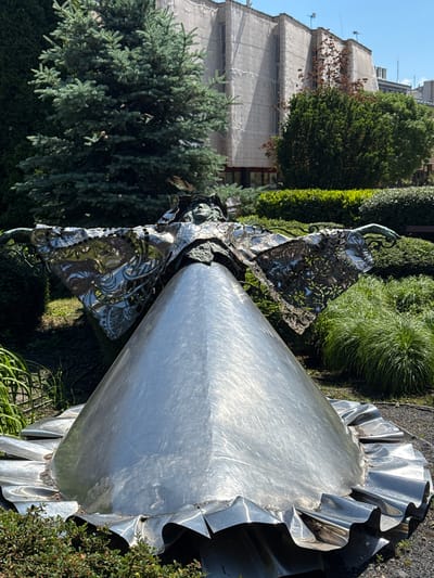 The image shows a stainless steel sculpture of a ballerina located outdoors in Siófok, Hungary. The sculpture is placed in a garden setting with various green plants and trees surrounding it. The figure of the ballerina is stylized, with a long, conical body and a skirt with ruffled edges. The upper part of the ballerina appears to be more detailed, with intricate metalwork forming the dress. In the background, a building is visible, partially covered by scaffolding and netting. The sky is clear.
