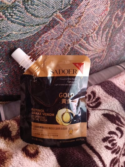 A gold and black pouch of Sadoer Retinol Snake Venom Peptide Facial Mask rests on a textured, colorful blanket. The pouch has a white plastic cap and a transparent window revealing a golden droplet graphic. Text on the pouch includes "SADOER", "RETINOL", "GOLD", "黃金", "RETINOL SNAKE VENOM PEPTIDE FACIAL MASK", and Chinese characters. The background is a soft, patterned blanket in shades of brown, beige, and white. The scene is indoors, and there is no indication of the city of Monastirische, Russia, or any people.