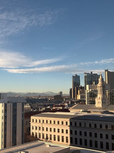 The image shows a cityscape against a clear blue sky with wispy clouds. Tall buildings dominate the skyline, ranging from modern skyscrapers to older, ornate structures. The buildings are densely packed together, creating an urban vista. Distant mountains are visible in the background, adding depth to the scene. The overall impression is of a vibrant, thriving urban environment with a backdrop of natural beauty.