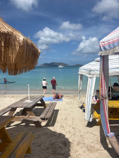 The image depicts a vibrant, sunny beach scene in Road Town, British Virgin Islands, under a clear blue sky with scattered white cumulus clouds. In the immediate foreground to the left, a portion of a light brown thatched beach umbrella is visible, alongside two wooden picnic tables; one is weathered brown, and the other, partially seen at the bottom left, is painted yellow. To the right, a striped beach cabana or tent, featuring white, blue, and red vertical stripes and fringed edges, encloses another yellow wooden picnic table and beach equipment.

On the light beige sandy beach, a male individual lies prone on a blue towel, facing away from the camera. Closer to the water's edge, a female wearing a white top, dark skirt, and white hat, holding a drink, walks towards the ocean alongside a male in a dark red t-shirt and dark shorts. In the clear, turquoise-hued ocean water, a person on a blue stand-up paddleboard is visible further left, while another individual wades closer to the shore. In the mid-distance, a white catamaran-style boat floats on the water, with multiple green, hilly islands forming the horizon in the background.