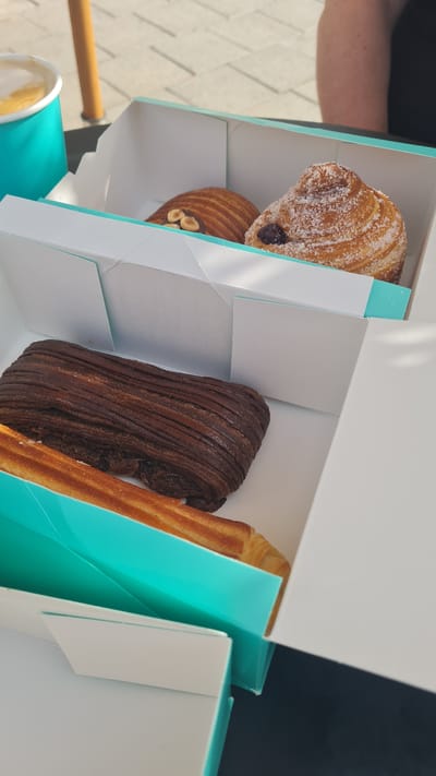 The image presents a top-down view of an open white and teal box containing a selection of various pastries, set outdoors in Kiryat Gat, Israel. The box holds at least four distinct baked goods.

In the upper left quadrant of the box, a golden-brown, flaky pastry, resembling a croissant or pain au chocolat, is visible. It is generously dusted with powdered sugar and contains a dark filling, likely chocolate or fruit jam. Directly below it, another golden-brown pastry with a similar texture is adorned with several small, chopped hazelnuts on its surface. Dominating the center of the box is a long, dark brown pastry characterized by prominent ridges, suggesting a rich chocolate-flavored croissant or similar item. To its right, a straight, golden-brown pastry, possibly a churro or a plain croissant stick, is neatly placed.

In the lower left corner of the frame, a teal paper cup, matching the box's accent color, is partially visible, holding a light-colored beverage that appears to be coffee or a latte. A light-colored wooden or metallic bar, possibly part of outdoor seating or a stroller, extends into the shot from the left.

The background reveals patterned light-colored paving stones, confirming an outdoor setting. Bright, natural sunlight illuminates the scene, indicating it is daytime, likely morning or early afternoon, under clear weather conditions. A person's arm or shoulder is faintly visible in the upper left background. The overall scene suggests a casual moment, perhaps enjoying a pastry and coffee break outdoors. No legible text is visible on any of the items.
