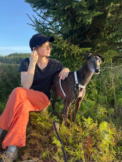 A woman wearing a black baseball cap, yellow-tinted glasses, a black t-shirt, and bright orange pants sits on a wooden post. She is looking off to the right of the frame, her right hand touching her ear. A brindle greyhound dog, wearing a white harness, stands beside her, facing the viewer. The two are in a grassy, outdoor area with a large evergreen tree on the right and a forested hillside in the background. The lighting suggests it is late afternoon or early evening. The image does not provide specific information to determine if the location is in the city of Hämeenlinna, Finland.