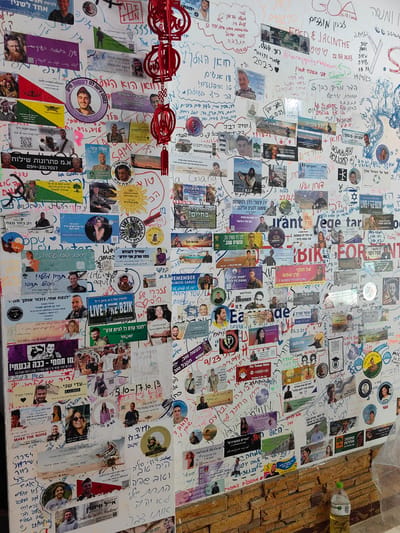 A cluttered wall in what appears to be a business establishment in Thành phố Hà Giang, Vietnam is densely covered with an assortment of photographs, memorial cards, and handwritten messages in Hebrew and English. The images primarily feature young people, many in military uniform, along with dates. Some of the images are memorial cards for fallen Israeli soldiers. The background wall is mostly white, covered in writing. Below the wall is a brick-like surface. A large bottle of liquid is near the lower right corner. Chinese-style hanging decorations are visible near the top of the wall.