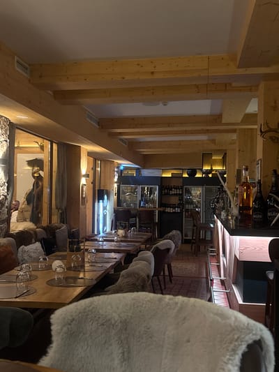 The image shows the interior of a restaurant or bar, likely located in Pamporovo, Bulgaria, given the ski resort atmosphere. The architecture features prominent wooden beams supporting the structure, typical of alpine-style buildings. A long table is set with glassware, suggesting a dining area, and chairs are draped with fur throws, adding a cozy, rustic ambiance. A bar area is visible in the background with bottles displayed on shelves, including recognizable brands like Belair. A display case with glassware and lighting is also noticeable. Through a window, a person and what appears to be a display of clothing are visible. The lighting is warm and inviting, highlighting the wooden elements and creating a comfortable environment.
