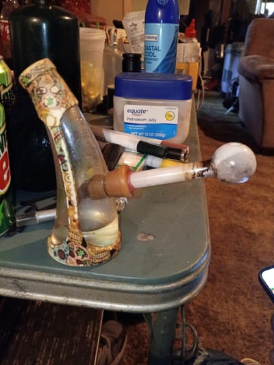 Here is a detailed description of the image:
**Scene Overview:** The image appears to be taken indoors. There's a collection of miscellaneous objects cluttered together on a surface. The focus is on what seems to be a used smoking device, surrounded by personal care products, pens, and soda.
**Subjects and Objects:**
* **Smoking Device:** There's a glass smoking device or bong present. It features a decorative pattern. The spherical bulb and stem seem to have residue suggesting it has been used.
* **Personal Care Products:** There's a blue can labeled "COASTAL COOL" which appears to be an air freshener. There is a jar of "equate Petroleum Jelly".
* **Other Objects:** There are pens, containers with liquid, a green and white soda bottle with a red label (likely Mountain Dew or a similar product), and a black cylinder.
**Environment:** The background appears to be the interior of a cluttered space, with a partial view of furniture. The surface on which the objects are placed appears to be a green or grey metal table or surface with an industrial look.
**Location:** The city of Clarksville, United States, is mentioned. However, the image itself does not offer visual clues to confirm that it was taken there. There are no clear local landmarks, business names, street signs, or other geographically specific visual elements within the frame.