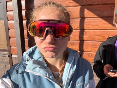 A woman with blonde hair and braids sits on a wooden bench, wearing iridescent ski goggles that reflect a cityscape. She is wearing a light blue and white jacket with "GLISSADE" visible on the left shoulder. Beside her, someone holds a smartphone. The bench is made of horizontal wooden planks. The reflection in her goggles suggests a city environment, and based on the location, this could be near Gory, Russia.