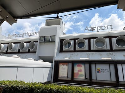 This is a photograph of the Lamezia Terme International Airport building in Lamezia Terme, Italy. The image is taken from a low angle, looking up at the facade of the airport. The building has a distinctive mid-century modern architectural style, with a white, curved upper section and a darker, glass-fronted lower section. Prominently displayed on the roof are the words "International" and "Airport" in large, silver lettering.

The upper part of the building features several circular windows, which appear to be ventilation openings, arranged in a row. The lower section has large windows displaying advertisements. One advertisement is for "caffè ARNONE" which states "The Calabrian Coffee Oak Wood Roasted Since 1953". Another displays "Candy & Co." with a website address. A third window shows the logo and text for "MARINA MILITARE Sportswear".

The sky is blue with scattered white clouds, suggesting it is daytime and the weather is fair. A line of lush green bushes is visible in the foreground, partially obscuring the base of the building. The overall impression is of a well-maintained, functional airport facility with a touch of retro charm. The presence of the "Marina Militare" advertisement suggests a connection to the Italian Navy, which may have a presence or a facility at or near the airport. The photograph captures a moment of ordinary activity at a transportation hub on a clear day.