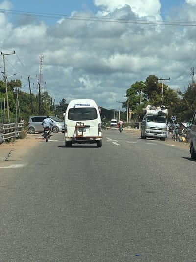 The image depicts a street scene in Muxara, Mozambique. The view is from a vehicle traveling on an asphalt road. In the foreground, a white minivan with a blue sign (partially readable as "ESOLACAO - MUXARA VIA VPP") is parked on the side of the road. A man is riding a motorcycle behind the van. A silver sedan is parked slightly ahead of the minivan. In the distance, another white van is visible, loaded with what appears to be cargo and surrounded by people. Trees line the roadside, and a tall communication tower is visible in the background, against a cloudy sky.