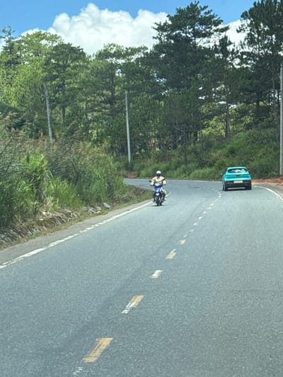 In Thôn 6, Vietnam, on a paved road, a person wearing a white helmet and a light-colored shirt is riding a blue and white motorcycle. The motorcycle is heading away from the camera, towards the right side of the frame. In front of the motorcycle, also heading away from the camera, is a teal colored car. The road is bordered by a grassy and vegetated embankment on the left and a forest of tall pine trees on the right. The sky is blue with scattered white clouds.