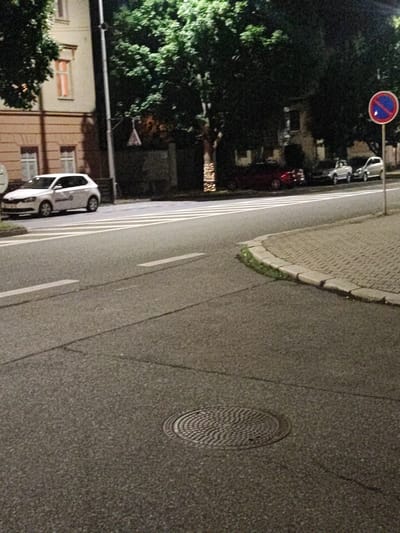 The image captures a street scene at night in Nový Jičín, Czechia. In the foreground, a dark asphalt road occupies the lower half of the frame, with a circular manhole cover visible on the right side. White road markings, including a pedestrian crossing, are present.
On the left side of the street, a white hatchback car is parked. Beyond the car, a building with light-colored facade and windows is visible. Trees with dense foliage line the side of the street and extend into the background.
In the background, several other cars are parked along the street. One car appears to be a red sedan, while others are lighter colored. A red and blue circular traffic sign with a blue diagonal line is mounted on a pole on the right side of the frame, indicating a parking restriction. The overall lighting suggests it is nighttime, with some artificial lights illuminating the scene.
