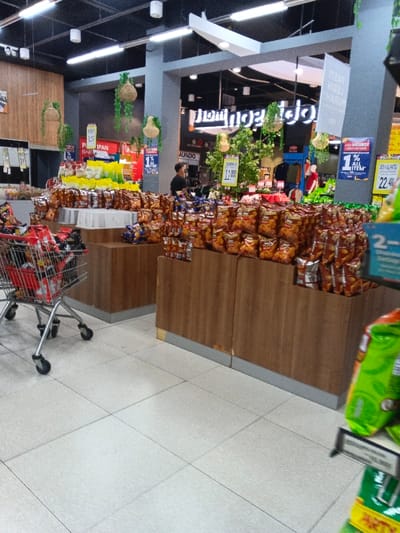 This image captures the interior of a brightly lit supermarket or grocery store, specifically focusing on a snack aisle. The scene is dominated by numerous bags of snacks, primarily potato chips or similar savory items, neatly displayed on tiered, wooden-clad stands that extend across the middle ground of the image. Some bags are also visible in a red metal shopping cart positioned on the left side of the frame.
In the background, a lone individual, a male wearing a dark shirt, stands near other display shelves. The store features a dark ceiling with modern white rectangular light fixtures and decorative elements like hanging plants in woven baskets. Signs are scattered throughout, indicating promotions and product information.
Visible text includes: "MUNDO" on a black sign; "SMX" and "11.AV" (likely a price like 11,000) on a smaller sign; "JADI MEMBER" (Become a Member) and "1% ALL ITEM" (1% off all items) on blue promotional banners; and "22,400" on a yellow sign. Other partial texts like "PI," "BAR," "IPAN," and "HARUM" are also visible. Upside-down text in the far background appears to include "MURAH," suggesting "cheap" or "affordable." The floor is tiled with light-colored, square tiles. The setting is likely in Sindanglaya, Indonesia, given the location context.