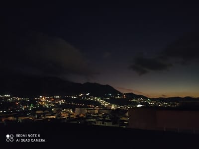 A night cityscape sprawls with illuminated buildings and streets stretching toward darkened mountains. Clouds obscure the sky above, hinting at a recent sunset or dawn. The lights of the urban area create a stark contrast against the dark silhouette of the hills, with a rooftop in the foreground.
