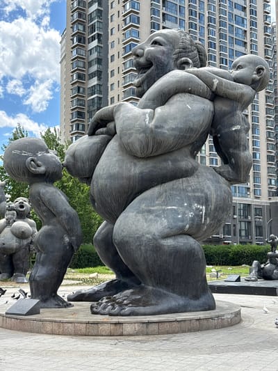 The image shows a bronze sculpture of people piled on top of each other. One person is on the ground, with other people sitting and climbing on them. In the background is a tall building with rows of windows. The sculpture is located in Changping District, China.