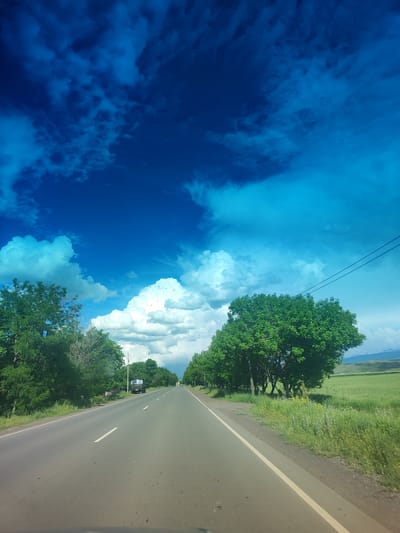 The image depicts a two-lane asphalt road stretching into the distance, bordered by lush green trees and fields. The sky is a vibrant blue with scattered white and darker blue clouds. A truck is visible on the right side of the road in the distance, and power lines traverse the upper right portion of the frame. The overall scene suggests a rural or semi-rural setting, likely during daylight hours. The location is identified as the city of Horom, Armenia.