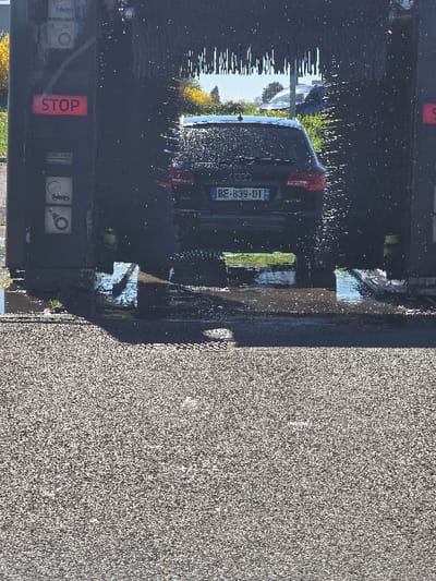 A dark-colored Audi station wagon (Avant model) with French license plate BE-839-DT is positioned within an active automatic car wash bay in France. The rear of the vehicle is covered in water droplets from the washing process, with visible water spray and wet brushes surrounding it. The car wash structure features a red illuminated "STOP" sign and instructional pictograms, including a Kärcher logo, on its left pillar. The ground inside the bay is wet with standing water, reflecting sunlight. The foreground is composed of dry, grey, granular pavement. Beyond the car wash, under bright daylight, green foliage with yellow flowers is visible, along with glimpses of other vehicles in the background. No people are present in the scene.