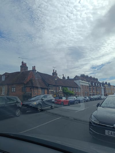 The image shows a street scene with parked cars and buildings. The buildings appear to be old, with tiled roofs and brick construction. There are several cars parked along the street, including a dark-colored Volvo SUV with the license plate "AK13 DYS" and a red convertible car. The sky is cloudy, and the overall impression is of a typical residential street in a historic town. The specific location is confirmed as Arun, United Kingdom, based on the visual cues and the license plate format which is consistent with UK plates.
