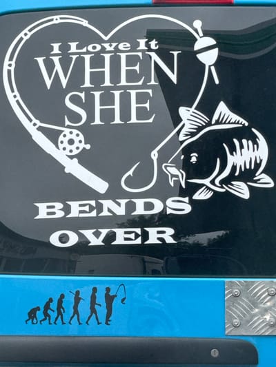 A white decal on the rear of a vehicle in South Oxfordshire, United Kingdom, features a fishing theme. At the top, a heart shape encloses the text "I Love It" above "WHEN SHE". Below this, the word "BENDS" is displayed, followed by "OVER". Within the heart, a fishing rod and reel are depicted, along with a fishing hook and a carp. To the right of the main decal, a depiction of human evolution is shown, progressing from an ape-like figure to a person holding a fishing rod with a fish. The vehicle itself is a bright blue.