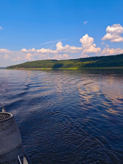 The image depicts a wide, dark blue river flowing towards a lush, green forested hill under a bright blue sky with scattered white clouds. The foreground shows a portion of a boat, including a metal grate and railing. The water has visible ripples and wakes, suggesting movement. The forested hill is dense with trees and rises gently from the riverbank. The sky is clear and bright, with the sun illuminating the clouds and casting a warm light on the scene, particularly reflecting on the water's surface. The location is the city of Podkamennaya Tunguska, Russia, and the overall scene captures a serene natural landscape from the perspective of someone on a boat.
