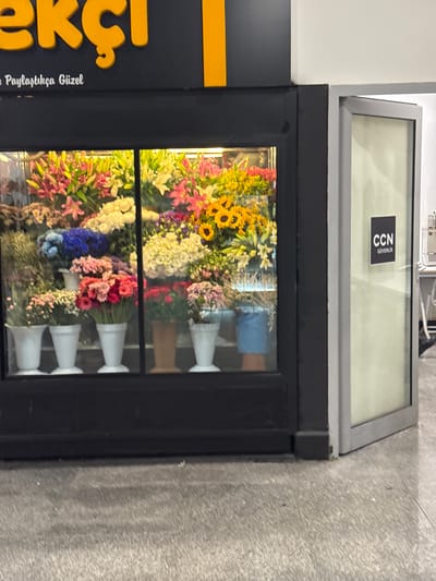 The image shows the exterior of a flower shop with a display window. The shop has a large yellow sign with Turkish text, partially visible. Below the sign, smaller white text reads "Paylaştıkça Güzel". The display window showcases a vibrant arrangement of various flowers, including pink lilies, yellow sunflowers, blue hydrangeas, and white chrysanthemums, housed in white and brown pots. To the right of the flower shop is a glass door with a dark square sign that reads "CCN GÜVENLİK". The floor is made of gray concrete. The scene appears to be in a commercial area.
