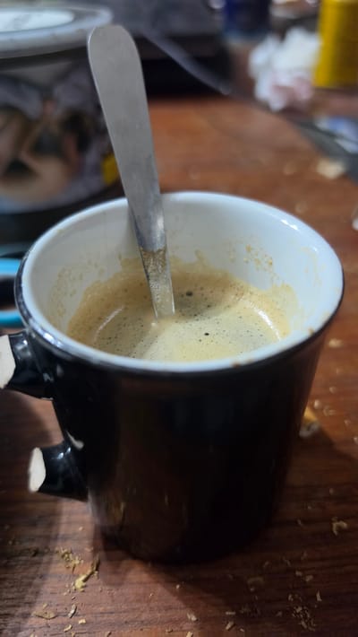 The image shows a close-up, angled view of a black coffee cup with a white interior, filled with a light brown beverage, likely coffee with crema. A silver spoon is partially submerged in the liquid. The cup rests on a wooden surface, which appears to have some debris scattered around it. In the blurred background on the left side of the image, there are indiscernible objects and a faint suggestion of a human figure. The location in Magny-les-Hameaux, France, cannot be determined from the image.