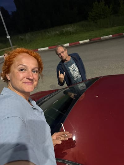 The image features two people near a red car at night. The person in the foreground is a woman with short, reddish-blonde curly hair and blue eyes, smiling at the camera. She is wearing a light blue collared shirt with a subtle textured pattern. Her right hand is visible, holding a lit cigarette. The background shows a man with short hair and glasses, wearing a dark blue denim jacket over a t-shirt with a graphic print. He is looking towards the camera and has his left hand raised, forming a "rock on" gesture with his fingers. The scene appears to be outdoors, possibly a roadside or parking area, with a dark grassy area and a road visible. A curb with red and white markings is also present. The lighting suggests it is nighttime. The overall mood seems casual and relaxed.
