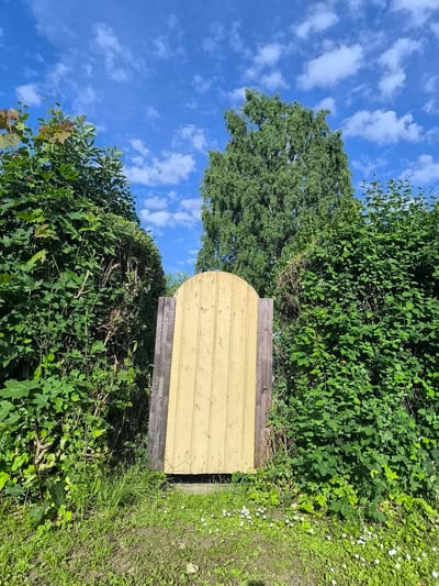 The image depicts a yellow wooden gate set within a dense green hedge. The gate has an arched top and is framed by rough-hewn wooden posts. Sunlight streams down, casting shadows and highlighting the vibrant green of the foliage. The sky above is a bright blue, dotted with fluffy white clouds, suggesting a clear and pleasant day. The ground in front of the gate is covered in green grass, with a scattering of small white wildflowers. In the background, a tall, leafy tree stands behind the hedge, its branches reaching towards the sky. The scene evokes a sense of a secluded garden or a hidden pathway, inviting exploration. The overall mood is serene and peaceful, with the natural elements creating a tranquil atmosphere. The image appears to be taken outdoors, likely during daytime, under fair weather conditions. No text is visible in the image. The location context provided is Rannamõisa, Estonia.