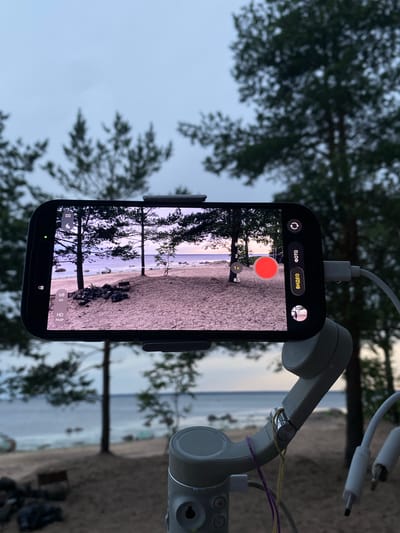 The image shows a smartphone mounted on a gimbal, capturing a video of a beach scene. The smartphone screen displays the camera interface with various settings such as "60 K/c" and "HD PA3P" visible. The scene being filmed includes trees, sand, rocks, and the sea under an overcast sky, suggesting a location in Primorsk, Russia, likely a coastal area. The gimbal itself is white and has cables connected to it. The overall atmosphere is calm and natural.