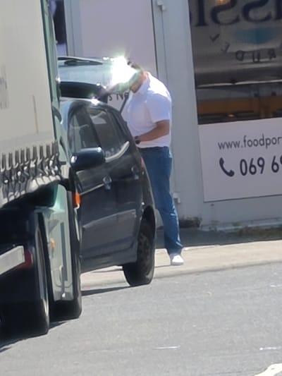 The image depicts a street scene in Zeppelinheim, Germany, on a sunny day. In the foreground, the side of a large white truck is visible on the left. To its right, a dark-colored car is parked, and a man is standing next to it. The man is wearing a white collared shirt and blue jeans. He is leaning into the car, with his head obscured by a bright glare. The background shows a building with a sign that includes the website "www.foodport" and a phone number "069 69". The overall environment appears to be an urban or commercial area.