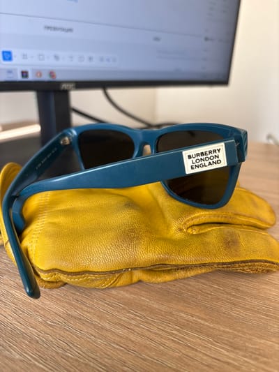The image depicts a close-up, angled view of a pair of blue-framed sunglasses resting on a single yellow leather glove, which lies on a light brown wooden desk surface. The sunglasses feature dark lenses and have a white rectangular label on the right temple arm displaying "BURBERRY LONDON ENGLAND" in black text. The leather glove appears worn, showing creases and darkened areas, particularly on the knuckles and stitching. In the blurred background, a black computer monitor, identified by a silver "AOC" logo on its base, is visible. The monitor screen displays what appears to be a word processing or presentation interface, with the word "ПРЕЗЕНТАЦИИ" (Russian for "Presentations") partially legible at the top. The scene is illuminated by an unseen light source, casting subtle shadows. This indoor setting is located in Зеленогорское сельское поселение, Ukraine.