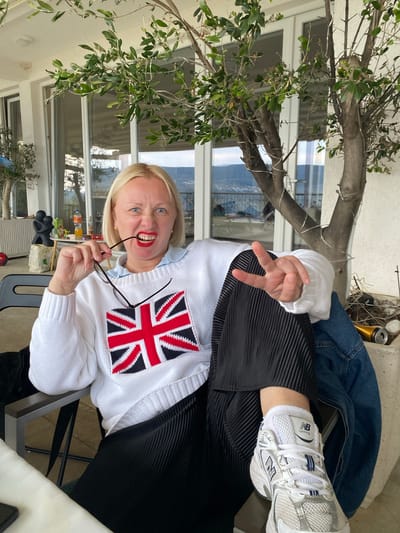 A middle-aged woman with blond hair is posing outside. She is wearing a white sweater with a Union Jack design, a black pleated skirt, and white New Balance sneakers. She is holding her glasses in her mouth and making a peace sign with her hand. The background includes a tree with green leaves, a building with large windows, and a glimpse of a cityscape, all consistent with the city of Trebesin, Montenegro.