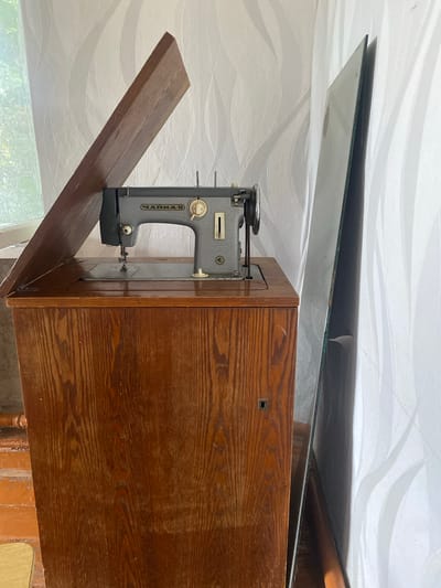 A vintage sewing machine, possibly a "Chaika-2," sits on top of a wooden cabinet in an interior room. The sewing machine is gray with some chrome accents and features Russian lettering. A wooden lid, propped open at an angle, partially covers the sewing machine and the top of the cabinet. The cabinet itself is made of dark, grainy wood and has a keyhole on its front. To the right of the cabinet, leaning against a textured, white wallpapered wall, is a large, flat pane of glass, possibly a mirror. The lighting suggests it is daytime, with light coming from a window on the left, which is partially obscured by a light-colored curtain. The floor appears to be made of wood or linoleum. The overall impression is of a somewhat cluttered, storage area or a room in an older home. The location is specified as Sakmara, Russia.