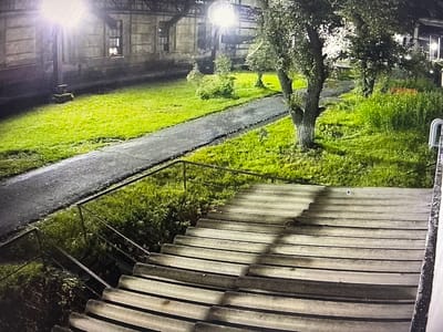 The image shows an outdoor staircase at night, likely in an urban or semi-urban setting. A bright light source, possibly a street lamp or floodlight, illuminates the scene from the left, casting long shadows. The staircase itself appears to be made of concrete or stone, with a metal handrail on the left side. The stairs are worn and uneven.
In the background, there is a paved pathway or road running alongside a grassy area. Several trees are visible, some with dense foliage. A building with windows is partially visible on the left. The overall impression is one of a utilitarian, perhaps slightly neglected, public space. There are no people or animals visible in the image.