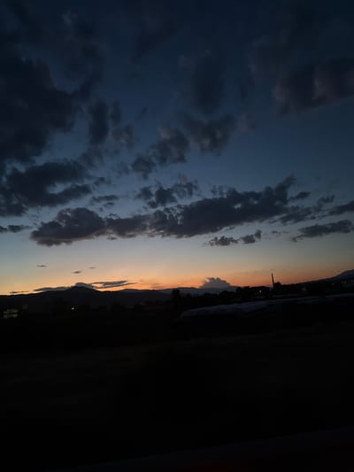 The image depicts a twilight scene in Novo Delchevo, Bulgaria. The sky is a deep blue, transitioning to a band of orange and yellow near the horizon. Dark, heavy clouds are scattered across the upper portion of the sky. Below the sky, silhouetted against the lighter colors of the sunset, are rolling hills and mountains. In the foreground, the landscape is dark and indistinct, showing the outlines of low buildings and possibly greenhouses or other agricultural structures. The overall impression is of a rural or semi-rural area on the outskirts of the town as day turns into night. There are no visible people or specific actions occurring in the scene.