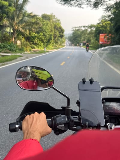 The image provides a first-person perspective from a motorcycle rider traversing a road in Vị Xuyên Commune, Vietnam. The foreground prominently features the right handlebar of the motorcycle, a hand in a red sleeve gripping the throttle, and a smartphone securely mounted vertically above the dashboard. A clear windscreen is also visible.
The road ahead is asphalt, marked with double yellow lines, and appears to stretch into the distance. Several other motorcycles or scooters can be faintly seen in the far background. Lush green trees and dense foliage line both sides of the road, suggesting a tropical or subtropical environment, with a distinct palm tree visible on the left. A blurry orange sign with unreadable text is partially visible through the trees on the right side of the road in the distance.
A rearview mirror, mounted to the handlebar, reflects two individuals. One person is wearing a yellow high-visibility vest, and the other appears to be wearing a red cap or helmet and a red jacket. These are likely passengers or other riders following behind.
Visible text includes the brand "SPIRIT BEAST" on a small metal component near the phone mount, and faint numbers such as "68" and "99" on the motorcycle's dashboard display, possibly indicating speed or mileage. The scene captures an active moment of motorcycling, suggesting travel or a tour on a clear day.