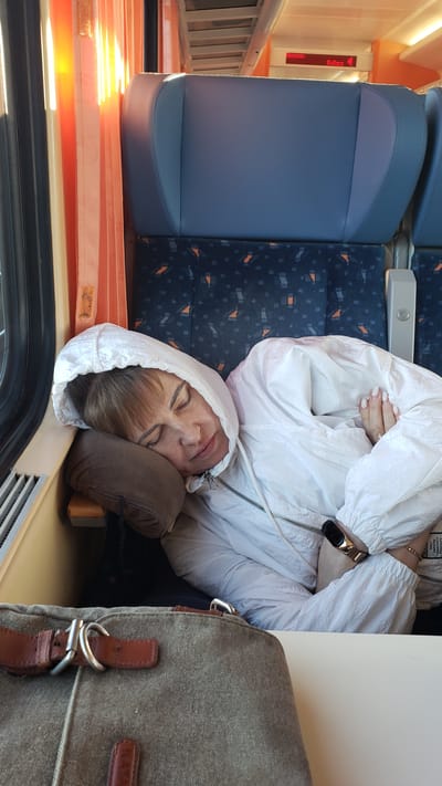 The image shows a woman asleep on a train, leaning her head on a brown pillow. She is wearing a white hooded jacket and her arms are crossed. A watch is visible on her left wrist. In the foreground, a brown canvas bag with leather straps is resting on a table. The train seats in the background are blue with a patterned fabric. The window shows orange curtains and sunlight streaming in, indicating it is daytime. Above the seats, a digital display shows "VÝCHOD: Košice 4". This train is likely traveling from Spišská Nová Ves, Slovakia, as indicated by the destination "Košice" on the display and the general context of the location.