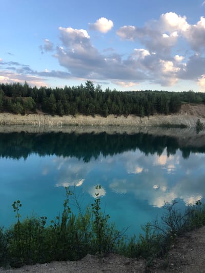 The image depicts a tranquil, reflective scene at a lake in Padlabenski rural council, Belarus. The clear turquoise water mirrors the sky, creating a symmetrical composition. On the left, a steep, light-colored cliff face is densely covered with dark green trees, suggesting a forest bordering the water. The cliff is eroded in a way that creates a textured, layered appearance.
The sky above is a mix of light blue and dramatic clouds, with patches of sunlight breaking through. The reflection in the water perfectly captures these elements, including the fluffy white clouds and the darker, more diffused clouds. The line where the land meets the water is sharp and defined, with the forest casting a dark, elongated shadow on the water's surface.
On the right side of the image, the bank of the lake is visible, covered in rough, sandy soil and dotted with sparse green vegetation and thin, bare branches of trees. The reflection here is less clear, showing the texture of the water and the dappled light.
The overall mood is serene and peaceful, with the stillness of the water enhancing the sense of calm. The lighting suggests it is either early morning or late afternoon, with the sun low in the sky. There is no visible text in the image.