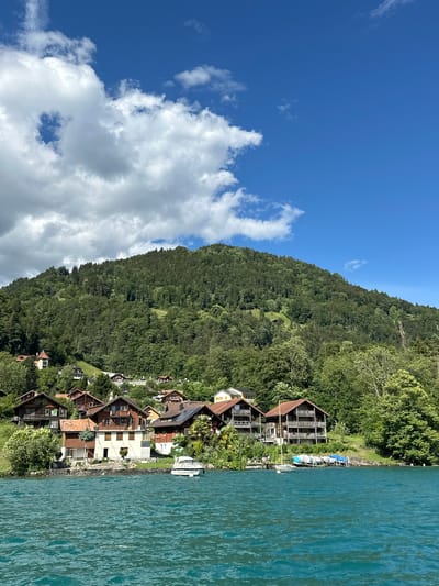 The image depicts a scenic view of Sundlauenen, Switzerland, on a clear day. In the foreground, a body of turquoise water, likely Lake Thun, stretches across the frame. Several boats, including a white motorboat and a sailboat, are moored near the shore. The middle ground features a line of houses with traditional Swiss architecture, nestled along the lakeside. These homes are characterized by their wooden balconies and pitched roofs, some adorned with solar panels. Behind the houses, a densely forested mountainside rises steeply, covered in a lush green canopy of trees. The sky above is a vibrant blue, dotted with fluffy white clouds. The overall scene conveys a sense of tranquility and natural beauty, characteristic of a lakeside village in the Swiss Alps.