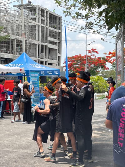 The image shows a group of people, likely participants in an event, gathered near a Pocari Sweat tent in บ้านสำนักตะแบก, Thailand. They are wearing black shirts with orange headbands. The event seems to be a "Tough Mudder" race, as indicated by the branding on the shirts. Some individuals are looking at their phones. In the background, there is a building under construction and a tree with red flowers. There are utility poles and overhead wires. Other individuals are seen in the background. The overall setting appears to be an outdoor event.